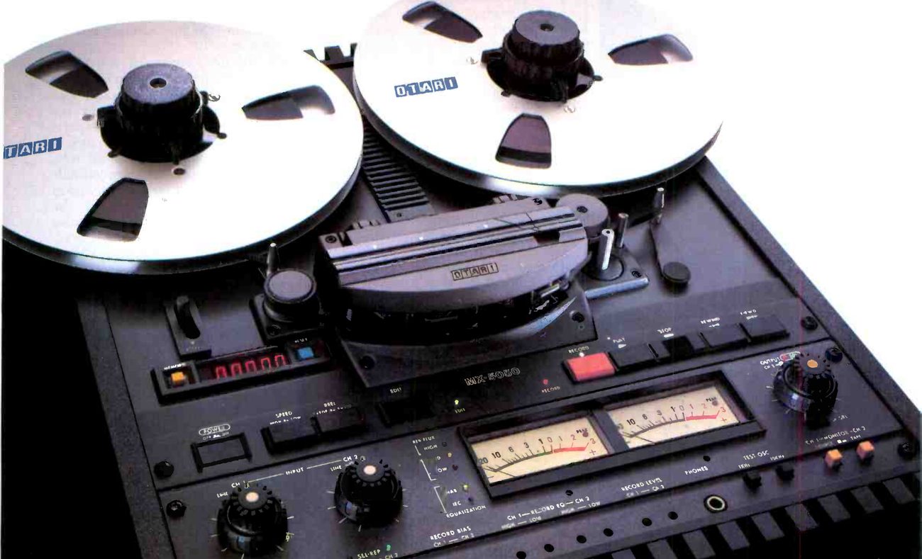 The MX-5050B-II photographed from above at an angle: two reels with OTARI on the flanges, the head assembly under its cover with an OTARI badge, the red LED tape timer at left, the MX-5050 escutcheon and a red record button, and the electronics panel below with two VU meters, input and output knobs and the record EQ and bias trims.