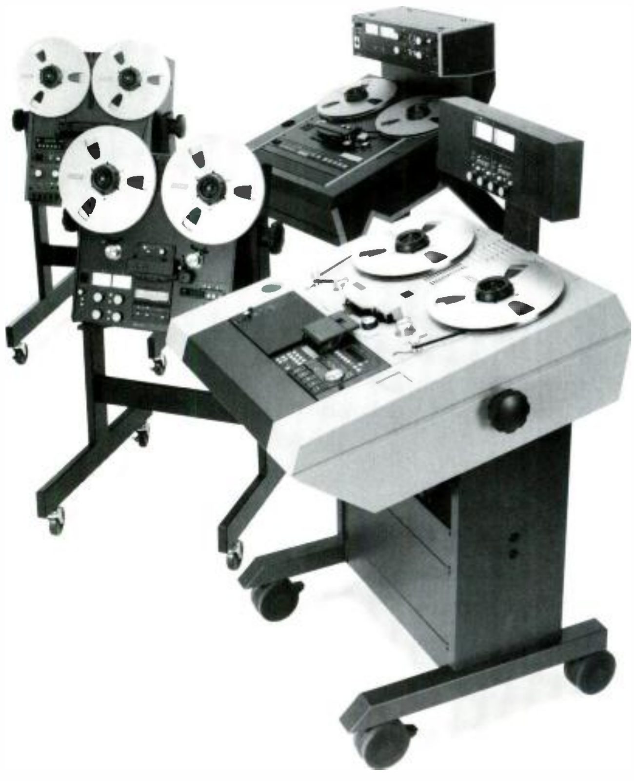 A group of Otari machines photographed together: two upright transports on wheeled stands at left, one behind the other, with ten-and-a-half-inch reels and their electronics below; a console machine with a raised meter panel behind; and in front a pale grey console transport with a large jog knob on its front edge, standing on a wheeled pedestal.