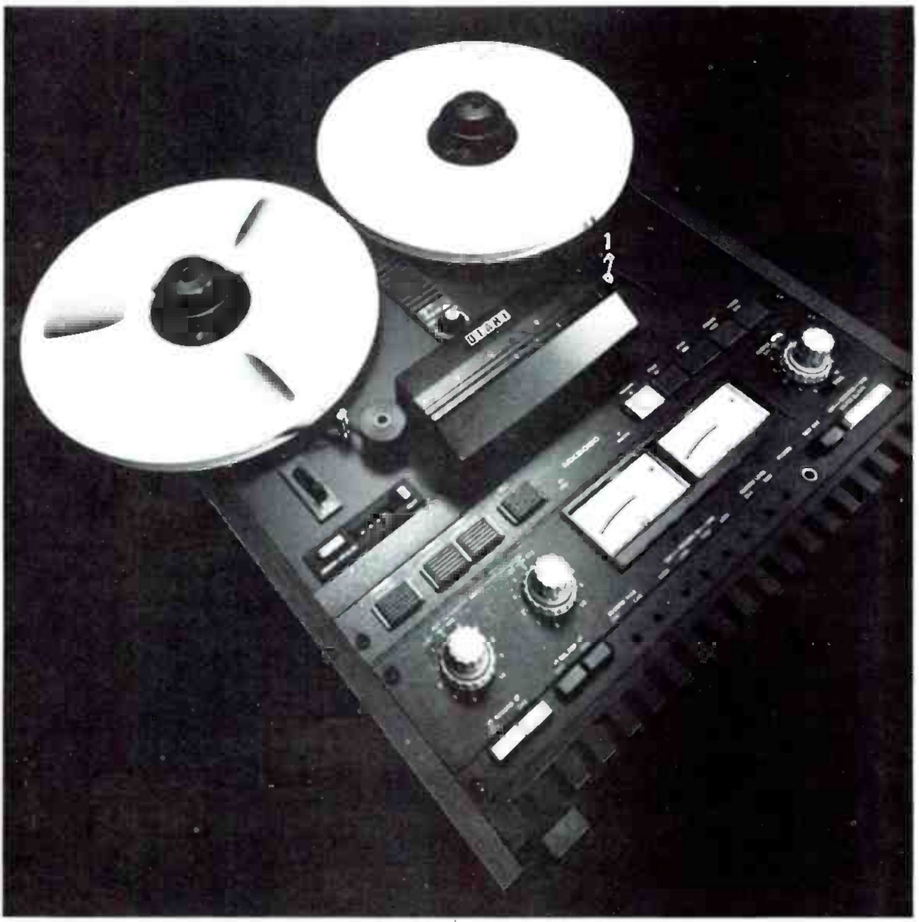 The MX-5050-B photographed from above, lying flat, with a reel on each hub: the head cover with the OTARI badge, the transport push buttons, the two VU meters, and the input and output level knobs on the angled electronics panel.
