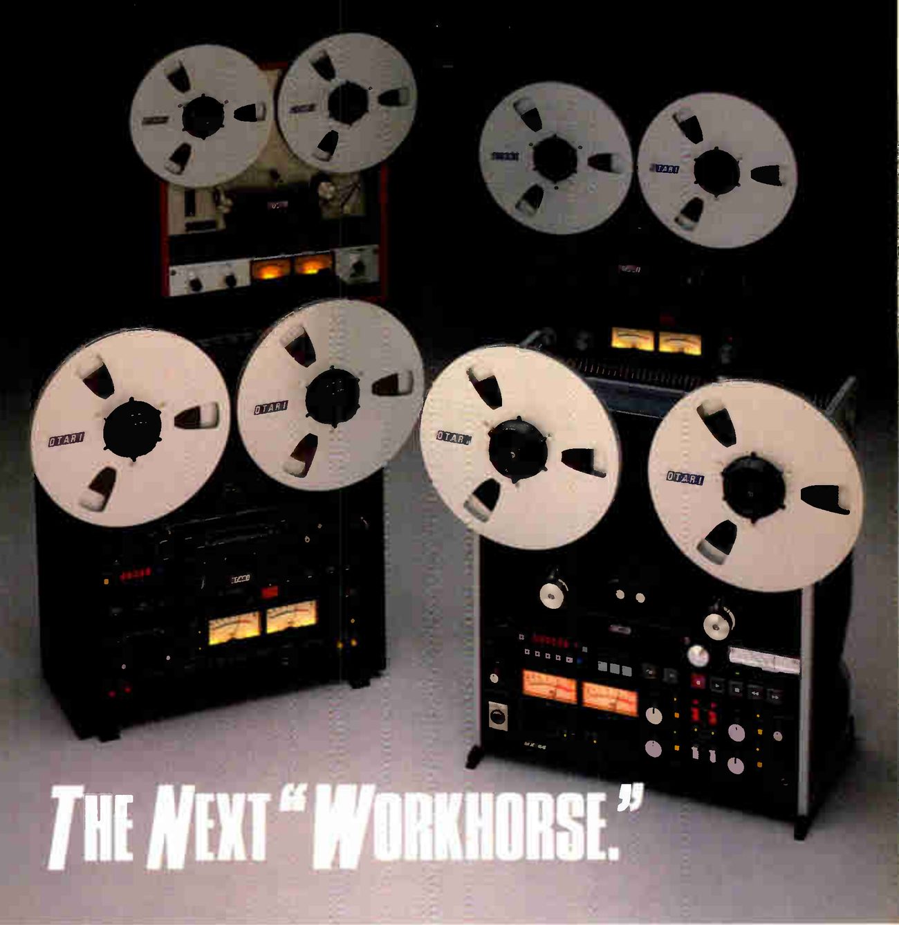 Four Otari two-channel recorders photographed in two staggered rows against a dark backdrop, OTARI on their reel flanges: at the back an older machine with red side cheeks and a second deck; in front, a BII-style machine with its red LED timer and a pair of lit VU meters, and a larger machine with rows of keys, lit meters and a small badge too blurred in the scan to read. The headline THE NEXT "WORKHORSE." is set in white across the floor in front of them.