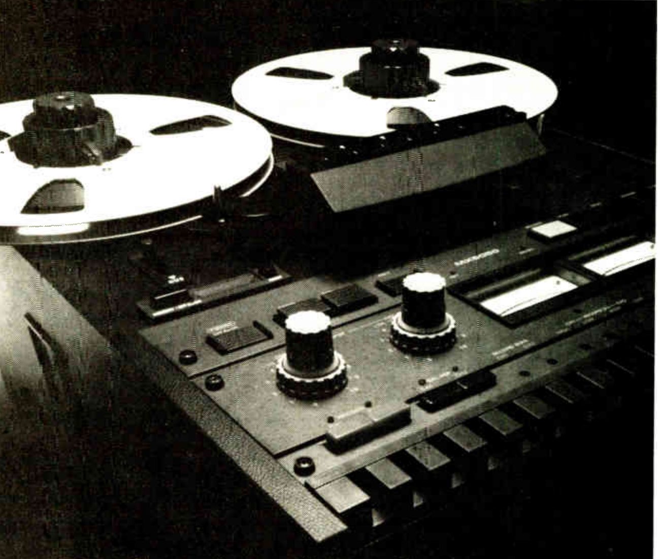 A low, close view across the MX-5050B's faceplate: two reels on the transport, the head cover, and in the foreground the electronics panel with its two large level knobs, meter windows and the row of transport keys along the bottom edge.