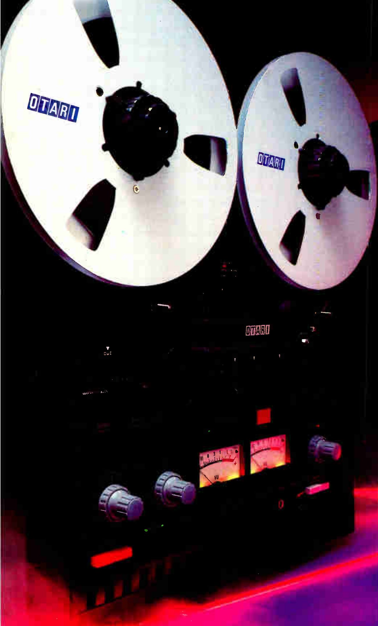 The MX-5050B shot low and close against a dark background lit red and violet: two large silver reels with OTARI hub badges fill the top of the frame, and below them the black faceplate with the OTARI badge, two lit VU meters and the level knobs.