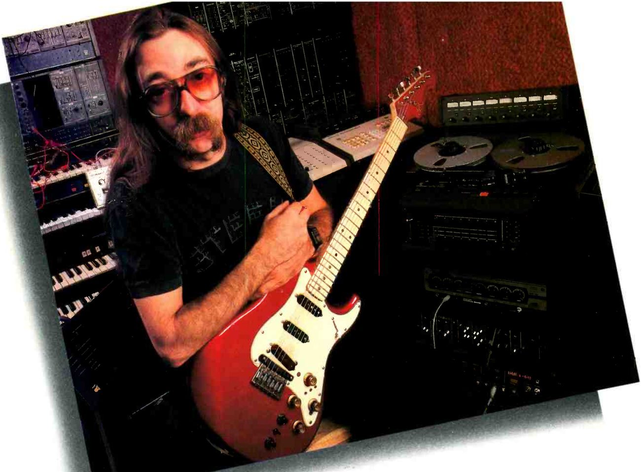 Jeff Baxter in his home studio holding a red electric guitar, synthesizer modules and keyboards behind him on the left, and on the right an eight-channel Otari with its meter bridge standing among racks of equipment.