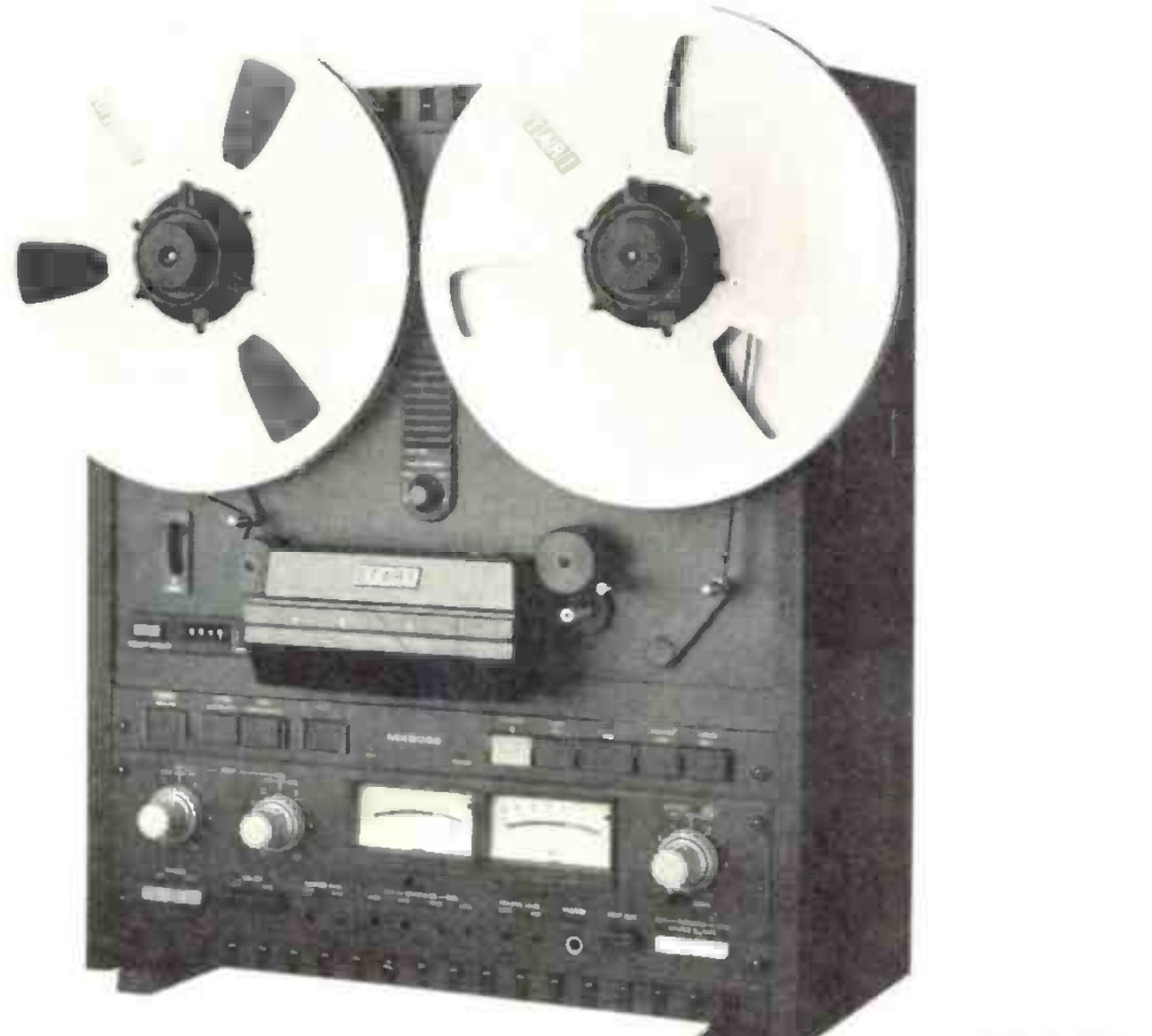 The MX-5050-B photographed from a slight three-quarter angle in its wooden side cheeks: two large reels on the transport, the head cover and tape lifter beneath them, the row of transport buttons, and the electronics panel with its two VU meters and level knobs.