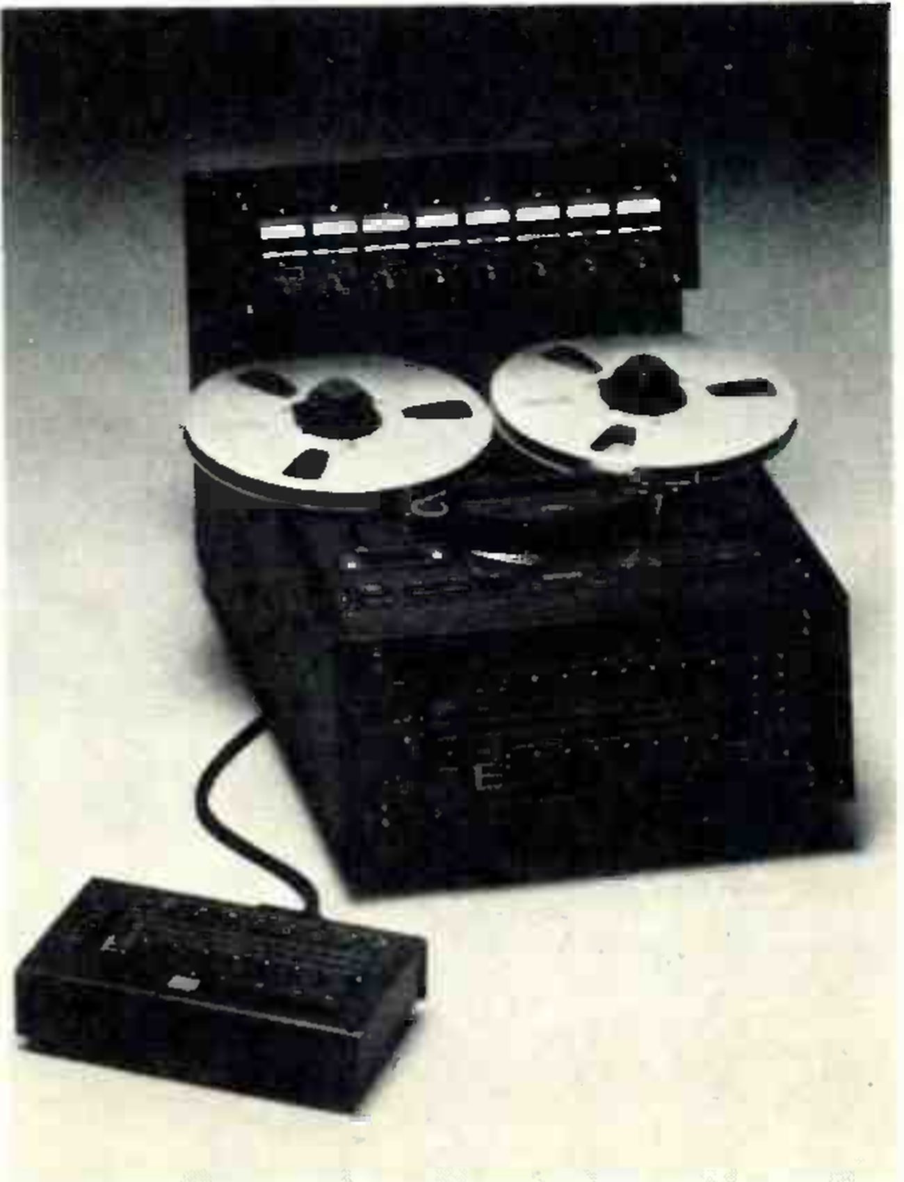The MARK III/8 photographed from a high angle on a pale ground: a raised meter bridge with eight illuminated meters across the back, two large reels on the flat transport, the dark sloping front panel below, and a remote control unit on a cable in the foreground.