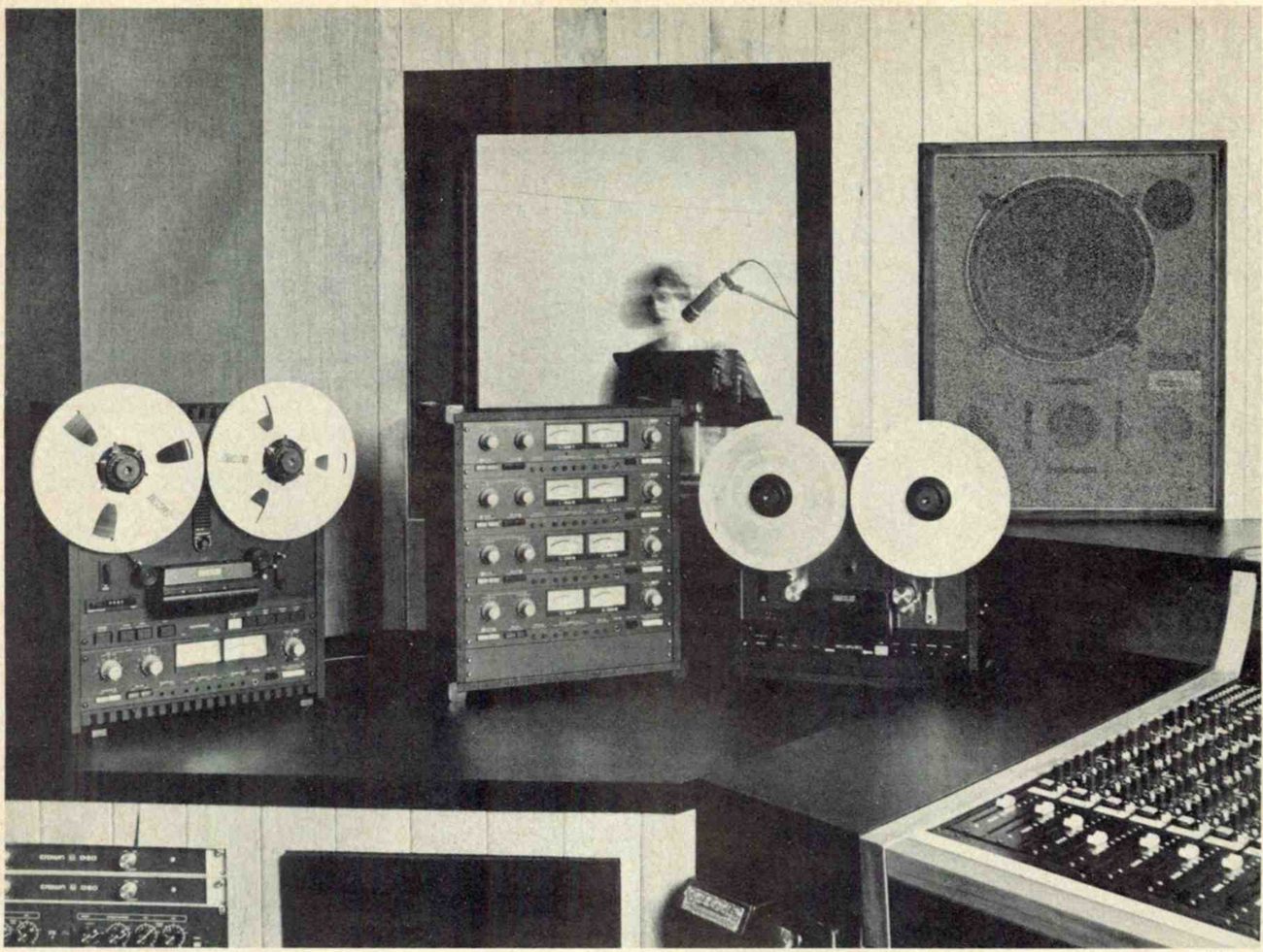 A studio control room photographed for the ad: at left a two-channel machine with two VU meters under slotted reels, in the middle an electronics bay with four rows of paired meters, at right a transport with solid-flanged reels, a monitor loudspeaker on the wall behind, a singer at a microphone seen through the studio window, and the corner of a mixing console in the foreground.
