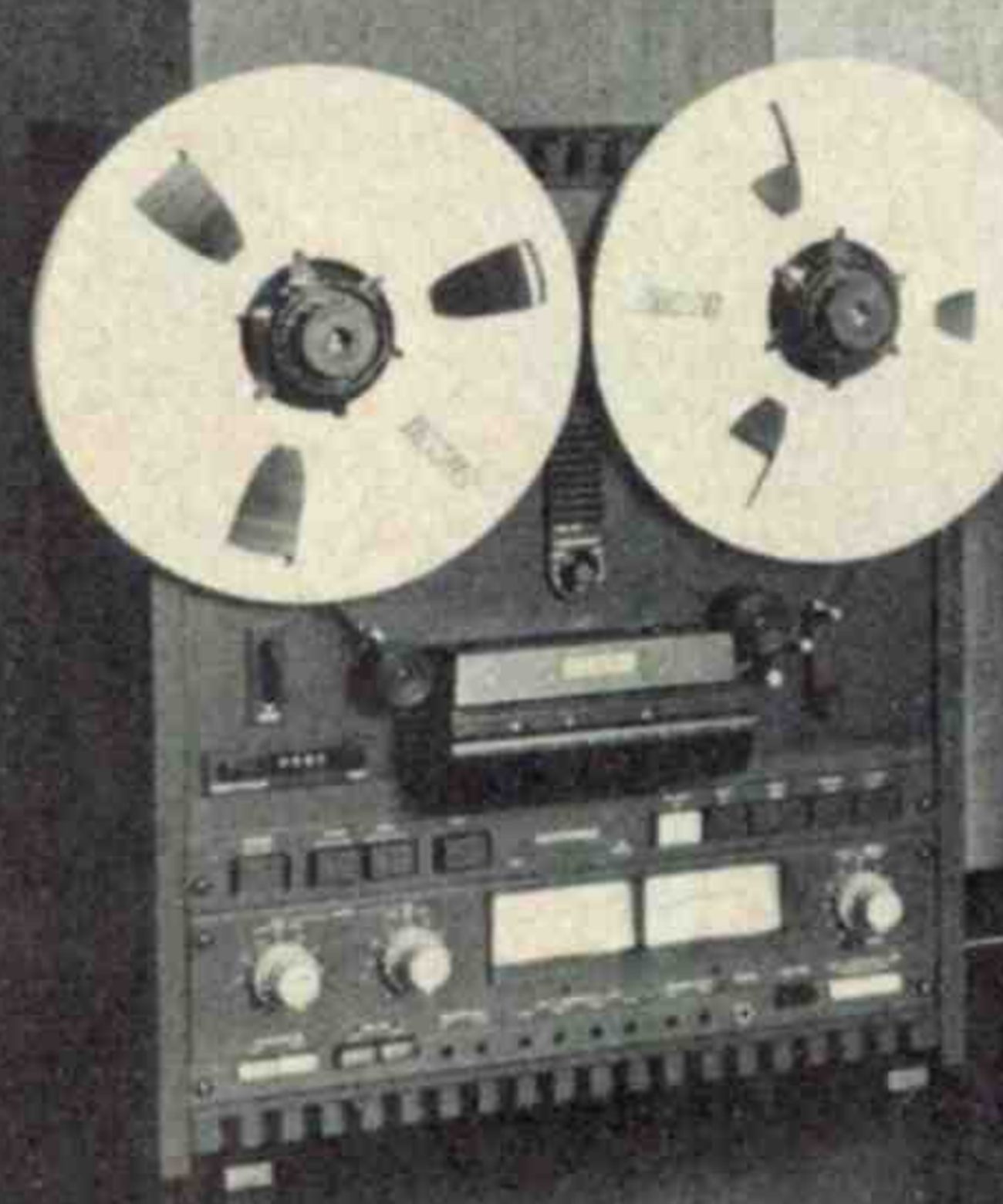 Detail of the left-hand machine: the reels, the head assembly under its cover, the transport keys, the two VU meters with level knobs either side, and the row of controls along the foot of the panel.