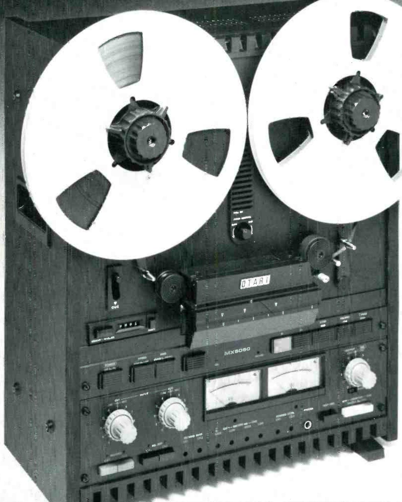 The MX5050B from a three-quarter angle in its wooden-sided case: two reels, the OTARI badge on the head cover, the CUE lever and tape counter at left, MX5050 on the electronics panel, and two VU meters between the input level knobs and the output knob.