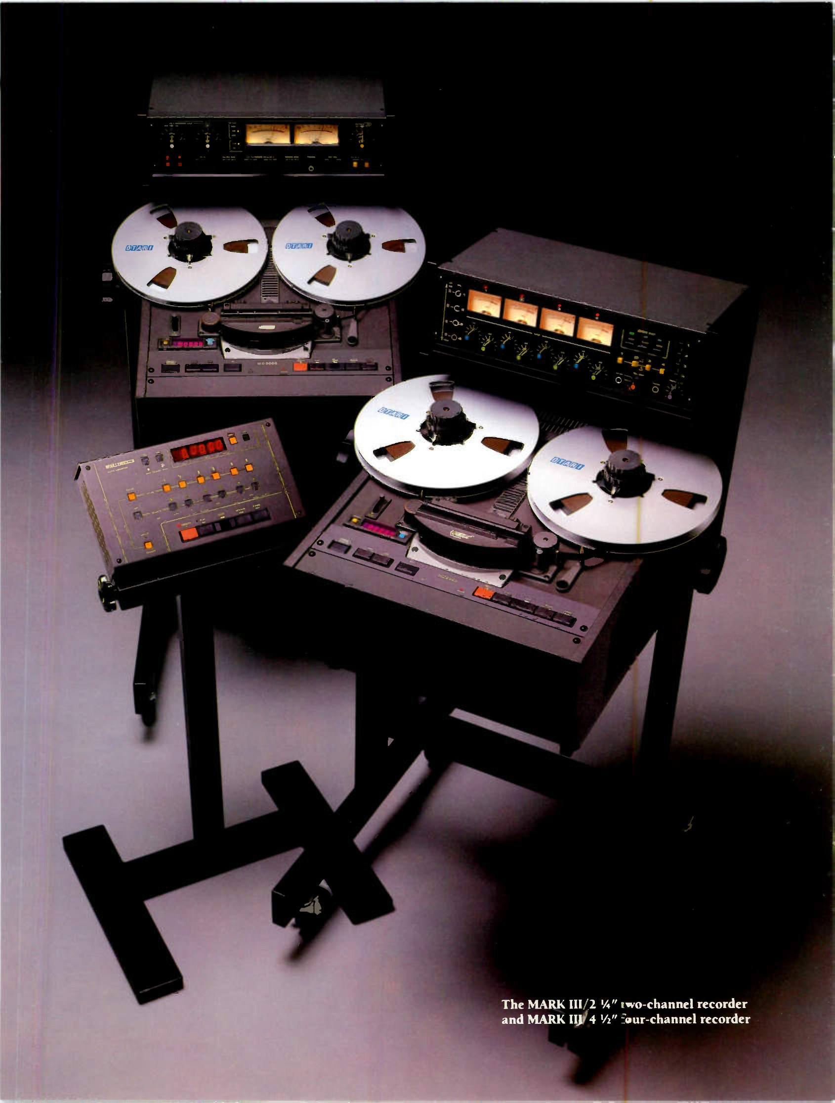 Two machines photographed together on floor stands in a dark studio: the MARK III/2 at the back with its rack-mounted two-channel meter unit above it, the MARK III/4 in front with a four-channel meter unit to its right, and an auto locator on its own stand at the left, all with white NAB reels fitted.