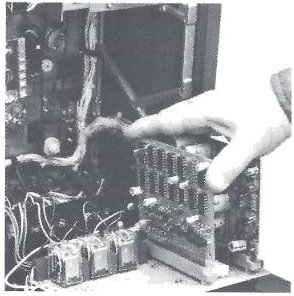 A hand drawing a transport logic board out of the MX-5050-8 transport from behind, with the rear panel lowered and the wiring and relays inside exposed