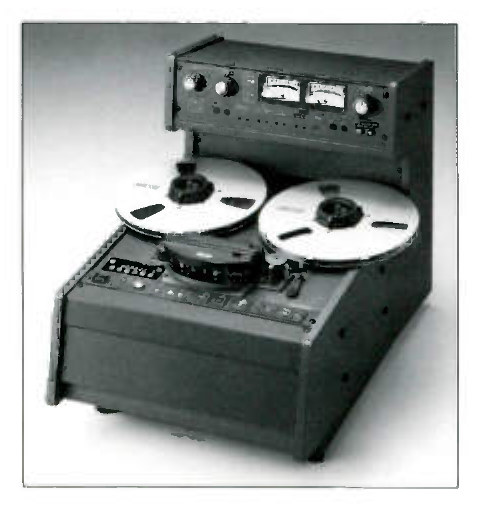 A recorder built as a desk-top unit, seen from the front and a little to the left: a separate electronics panel with two rotary controls, two lighted VU meters and a further knob stands across the back on a raised bridge, the transport lies back below it with two white Otari NAB reels flat and the head block between them, and the whole sits in a plain wedge-shaped cabinet on small feet.