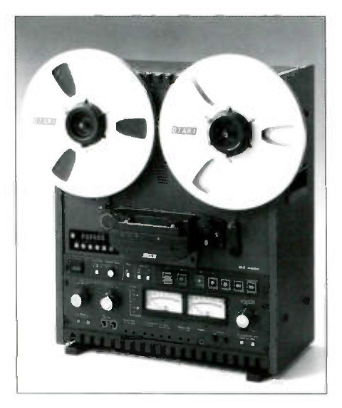 A recorder standing upright, photographed slightly from the left against a grey ground: two white Otari NAB reels side by side at the top, the head block and tape path below them behind a hinged cover, a row of six small keys at the left of the deck, and across the lower panel a pair of large knobs, two lighted VU meters, four square function keys and a rotary control at the right, over a finned vent strip along the bottom edge.