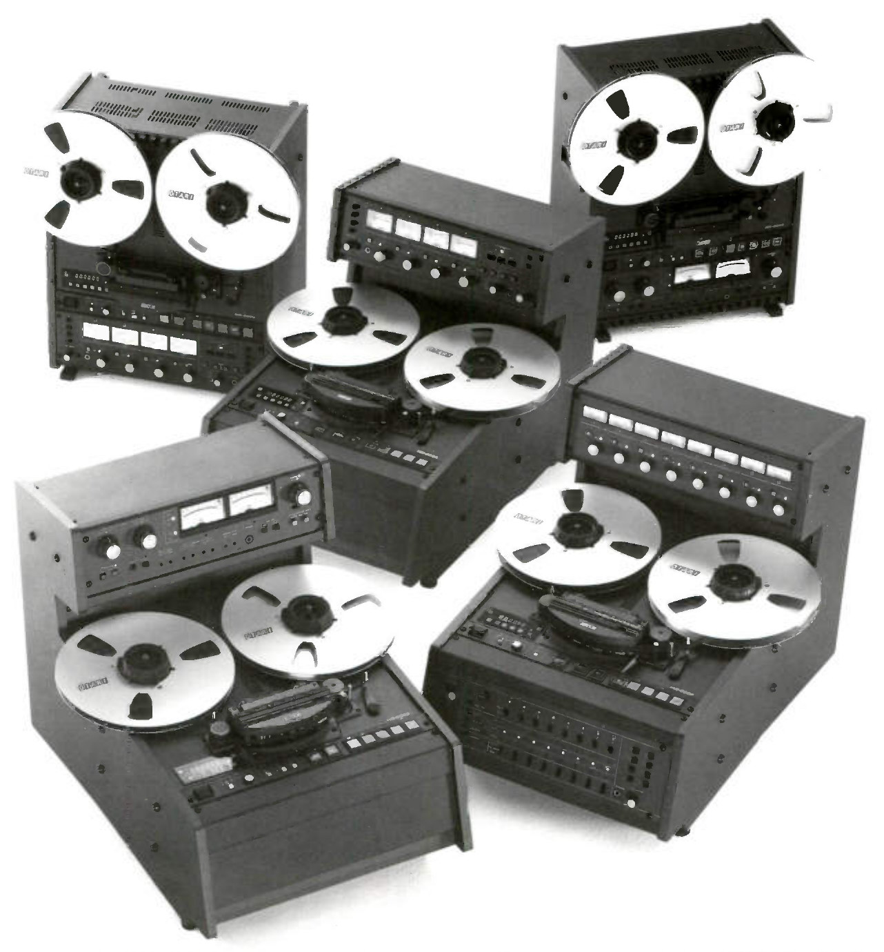 Five machines of the series grouped together on plain white paper, each carrying a pair of large pale reels. Two at the back stand upright, their reels vertical above the transport and electronics in the cabinet below; the three in front are desk-top cabinets whose decks lie back, so their reels are seen from above, each with its electronics on a raised bridge behind the deck. The bridge of the desk-top at the front left carries two meter faces; the bridges of the desk-top in the middle and of the larger one at the front right each carry a row of meter faces, the latter over a matching row of knobs.